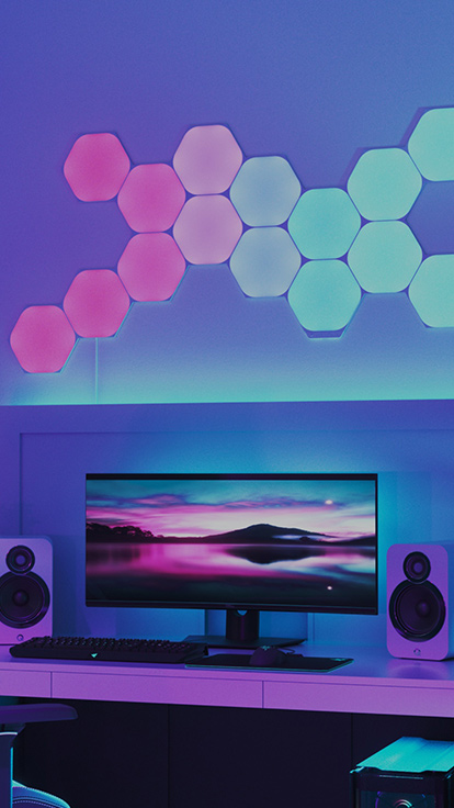 This is an image of Nanoleaf Shapes Hexagons on a wall above a monitor on a desk. They are RGB lights that are perfect for the gamer in your home environment. The modular color changing light panels connect together with linkers and have over 16 million colors.