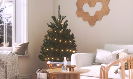 This is an image of a 10 panel layout of Nanoleaf Elements Wood Look Hexagons mounted on the wall beside a couch and next to a Christmas tree in the living room. Modular smart light panels that are secured to the walls with included adhesives and connected together with linkers to create a Heart shaped design. The wood look hexagons are the perfect decorative piece that adds style and personality in the living room while doubling as a customizable ambient light.