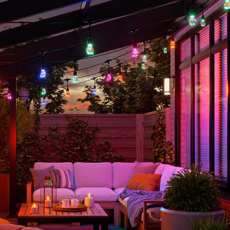Outdoor patio with multicolor outdoor string lights, hanging over a cream-colored corner couch with pillows and a striped blanket. Next to the couch is a plant by a small table with candles and a tray holding two mugs. Behind the couch, a wooden fence with creeping vines and a window emitting a pink and purple glow.